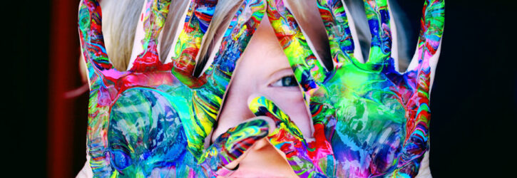 Child holding hands up in front of face. Hands are covered in rainbow paint