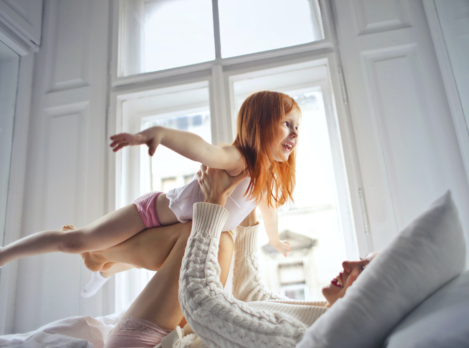 Photo of a mother ona bed holding her daughter above her in a flying position