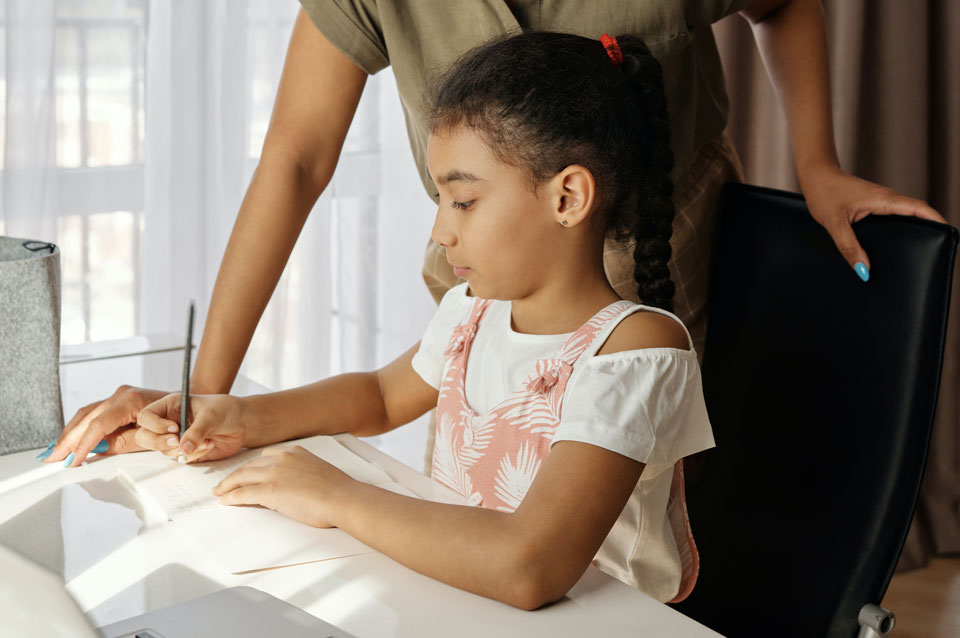 Child writing homework with mother standing next to her to help