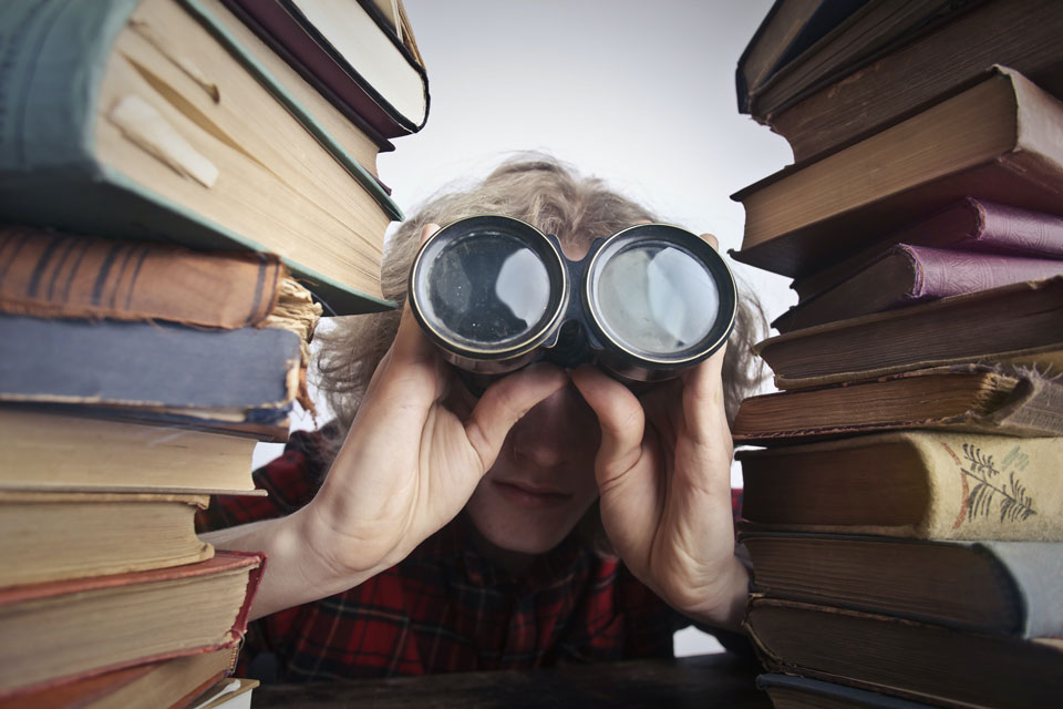 person staring through binoculars surrounded by piles of books