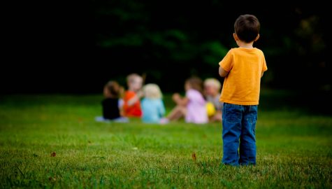 Young child standing isolated from a group playing on grass in front of him