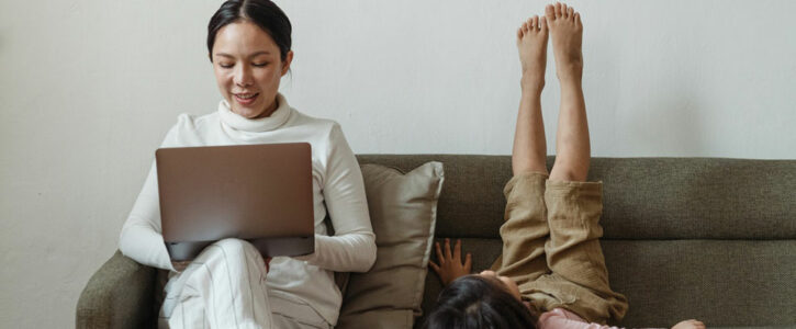 Mother sitting on a sofa looking at her laptop, child next to her is lying on sofa with feet in the air