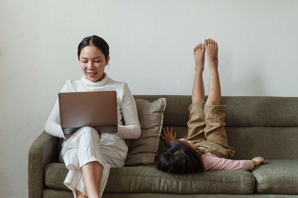 Mother sitting on a sofa looking at her laptop, child next to her is lying on sofa with feet in the air