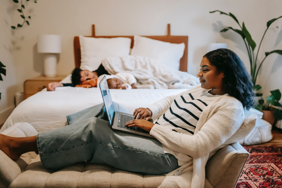 mother on laptop, father and child asleep on bed behind her