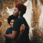 250228 Mental Health Bundle. Unsplash licence free by eye-for-ebony A parent and child embrace in front of a wall. The parent is shown in side profile, while the child faces slightly more toward the camera. Both have their eyes closed and wear soft smiles. Unsplash license-free image by eye-for-ebony.